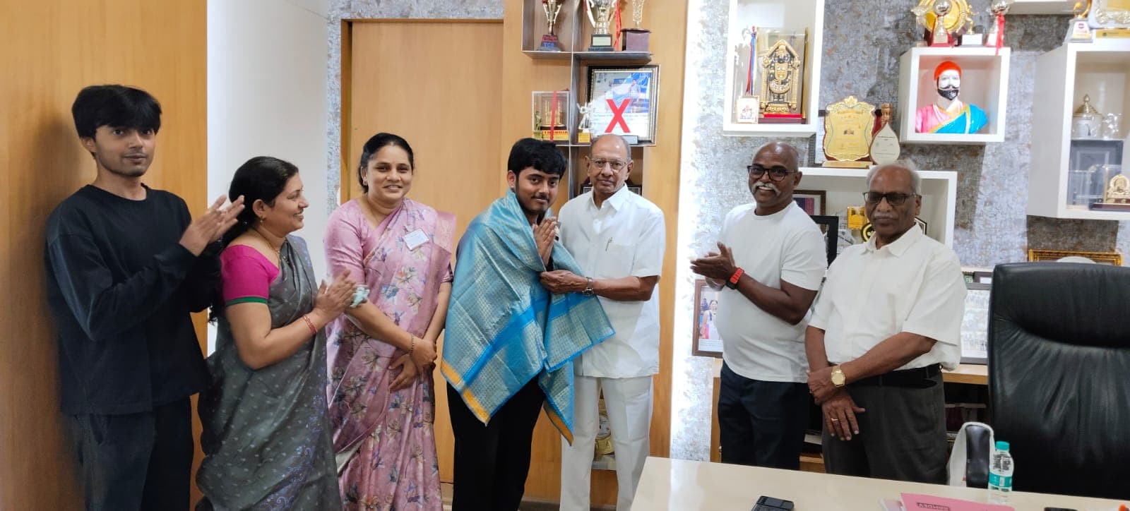 A son being felicitated with a ceremonial shawl by officials in a trophy-lined office