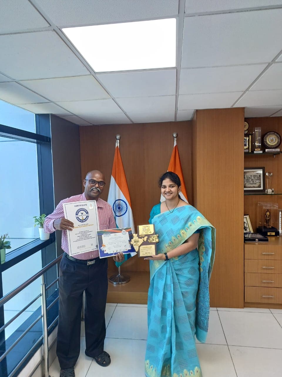 Bobba Ravinder Reddy and a colleague holding his International Records of Excellence certificate and plaque in front of Indian flags