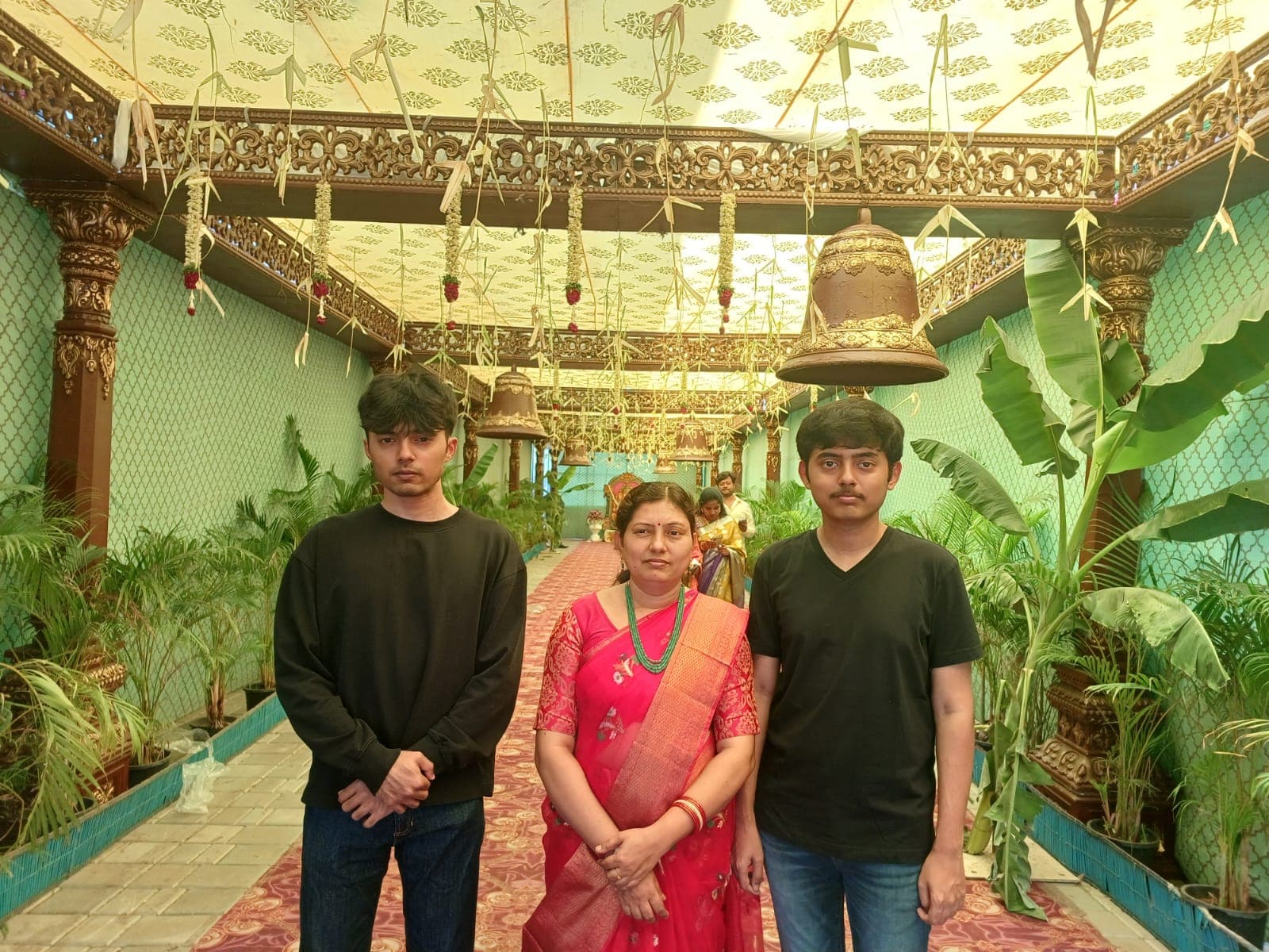 Two sons and their mother in a temple corridor lined with hanging bells and floral garlands