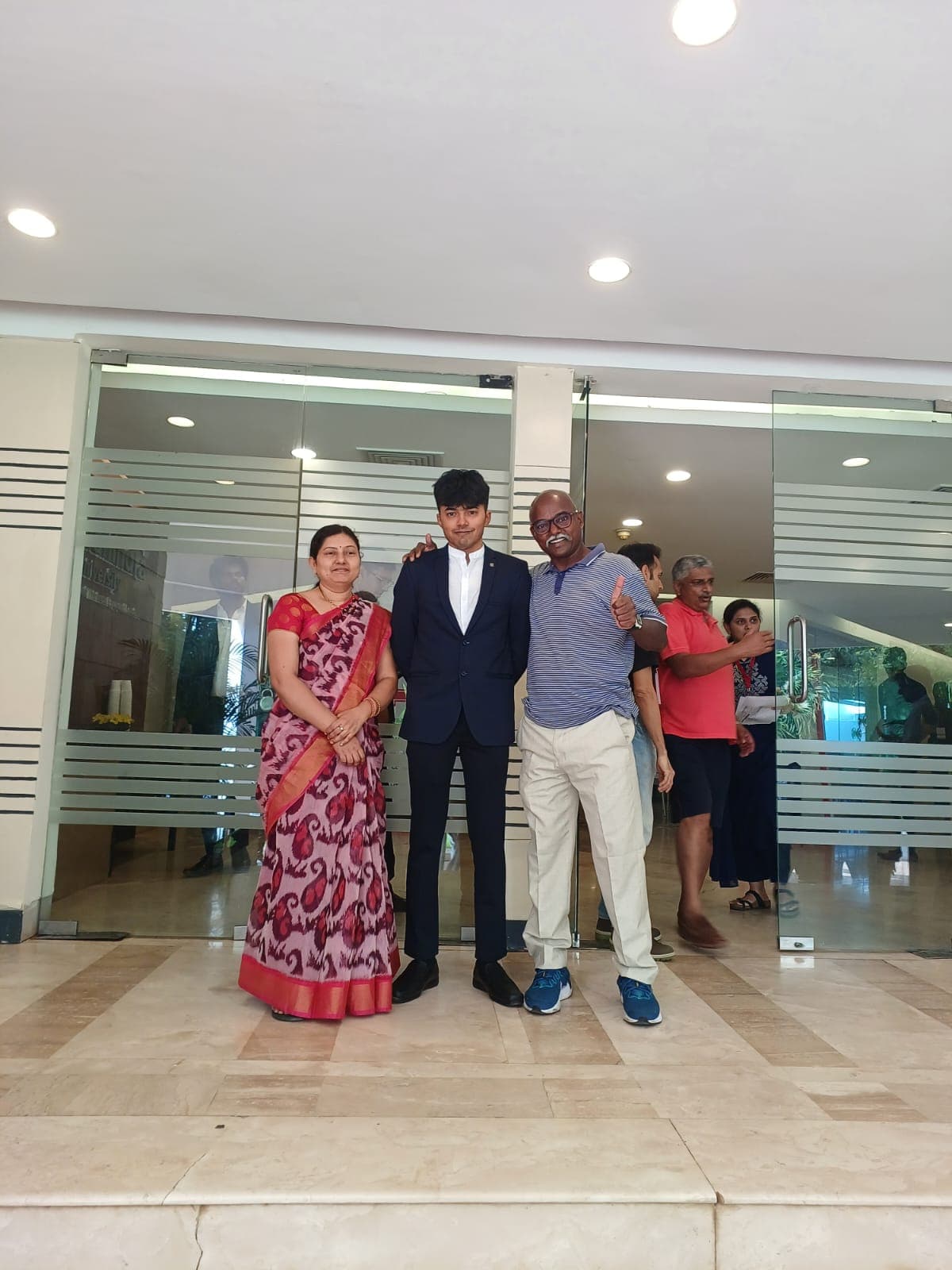 Bobba Ravinder Reddy and his wife posing with their son outside a building entrance, giving a thumbs up
