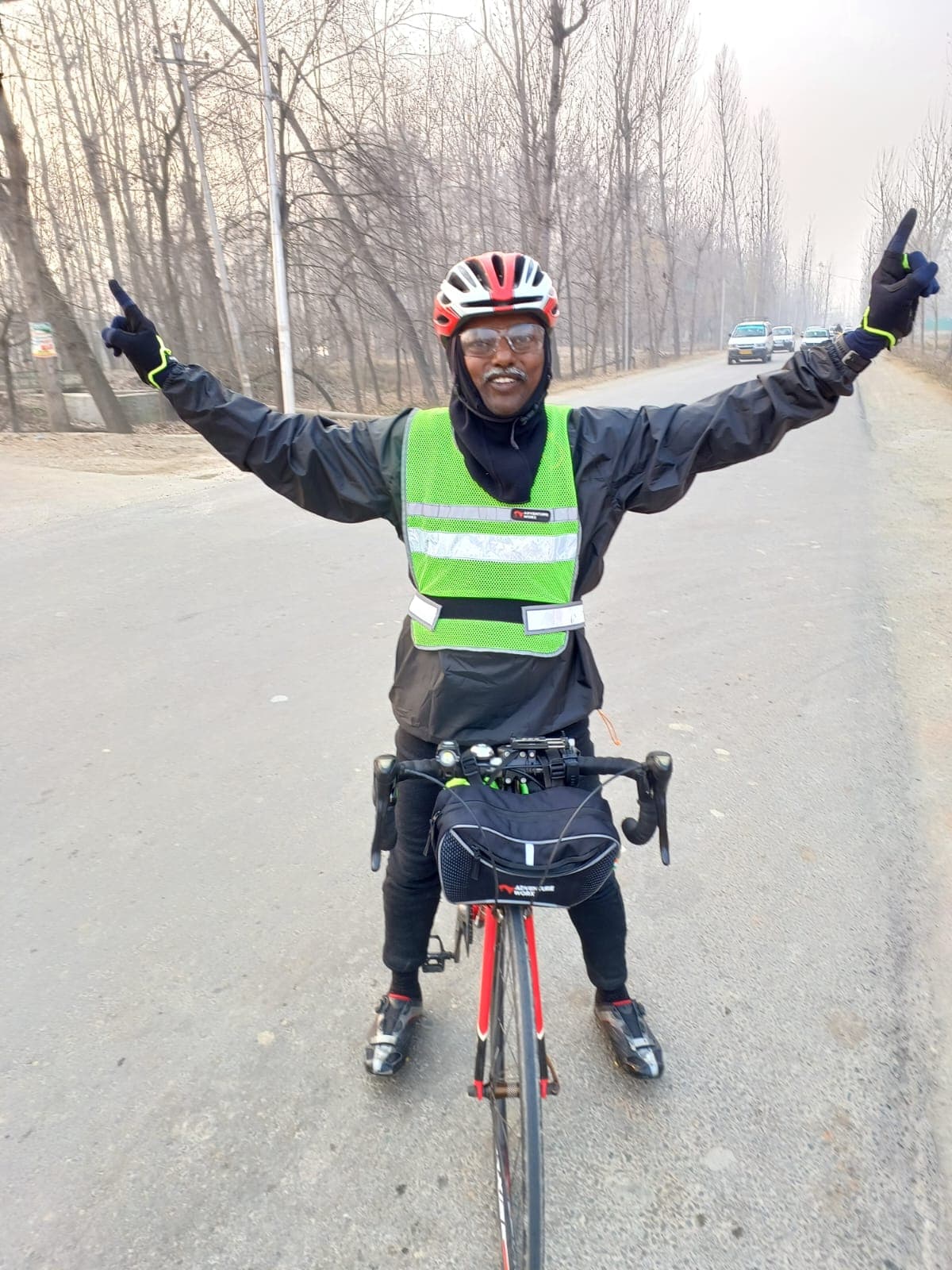 Bobba Ravinder Reddy raising both arms in celebration while cycling down a tree-lined winter road